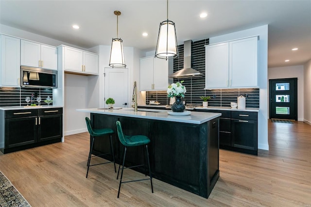 kitchen featuring white cabinetry, wall chimney range hood, pendant lighting, a center island with sink, and light wood-type flooring