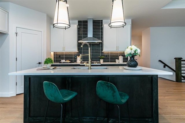 kitchen featuring an island with sink, hanging light fixtures, wall chimney range hood, and light hardwood / wood-style floors