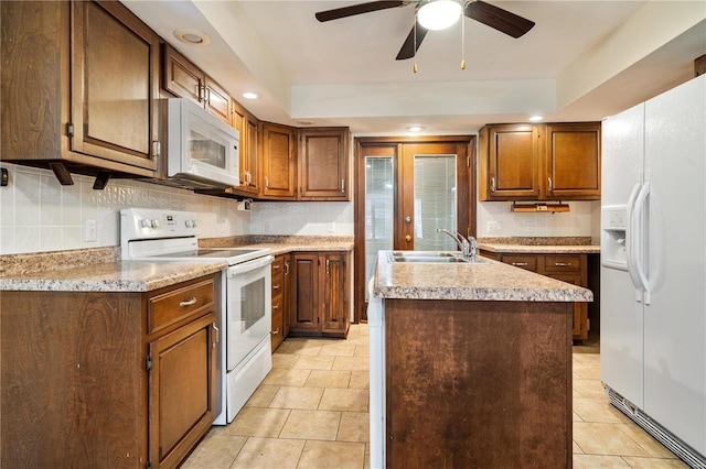 kitchen featuring white appliances, a kitchen island with sink, sink, ceiling fan, and a tray ceiling