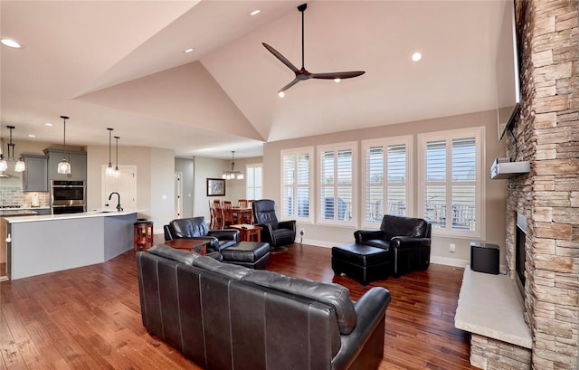 living area with high vaulted ceiling, a stone fireplace, wood finished floors, and ceiling fan with notable chandelier