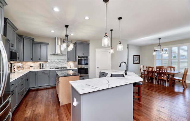 kitchen with a center island with sink, a sink, gray cabinetry, appliances with stainless steel finishes, and wall chimney exhaust hood