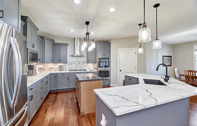 kitchen with a kitchen island with sink, gray cabinets, stainless steel appliances, wall chimney exhaust hood, and a sink