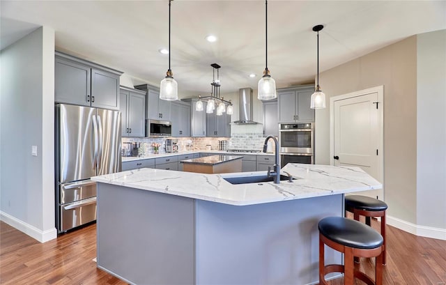 kitchen featuring wall chimney range hood, an island with sink, gray cabinets, and stainless steel appliances