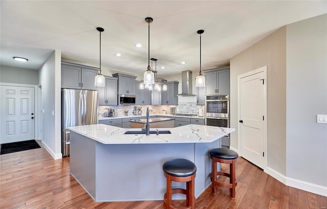 kitchen featuring a large island, gray cabinets, a sink, stainless steel appliances, and wall chimney range hood