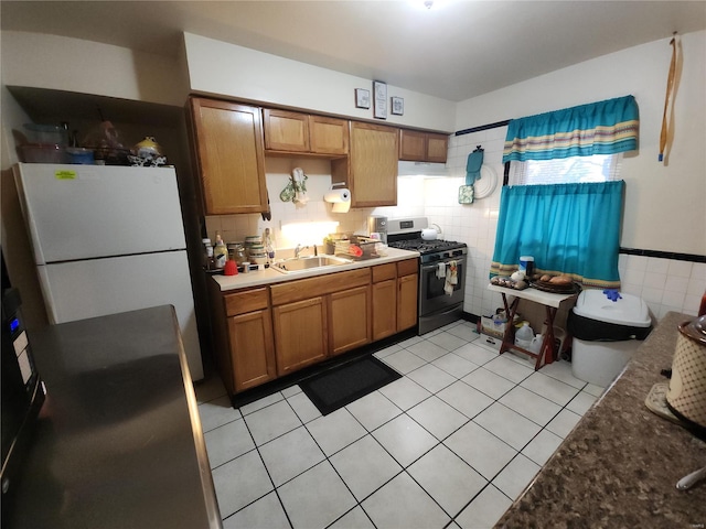 kitchen featuring tile walls, brown cabinetry, freestanding refrigerator, a sink, and gas range