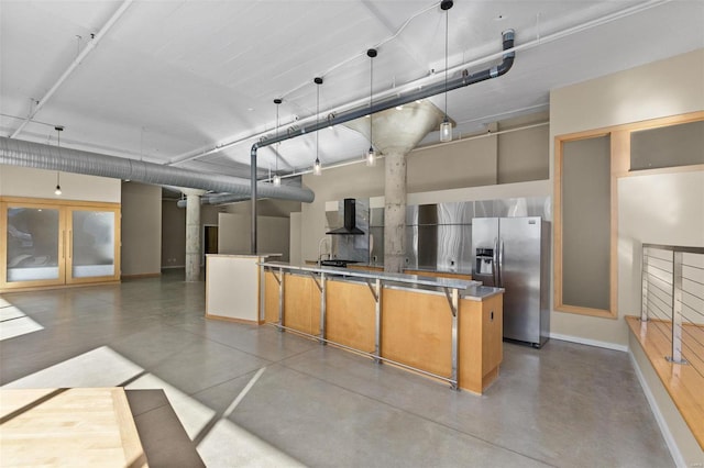 kitchen featuring hanging light fixtures, wall chimney exhaust hood, concrete flooring, stainless steel counters, and stainless steel fridge with ice dispenser