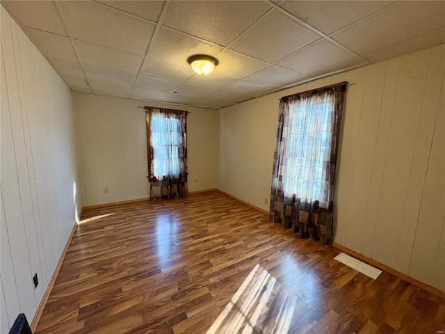 empty room with wooden walls, hardwood / wood-style floors, a drop ceiling, and plenty of natural light