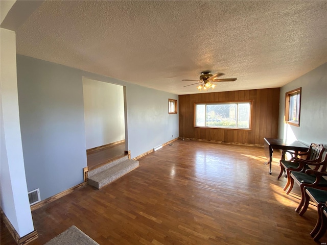 unfurnished living room with a textured ceiling, ceiling fan, dark wood-type flooring, and wooden walls