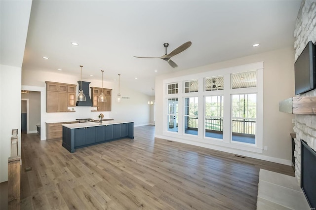 kitchen with a kitchen island with sink, a stone fireplace, hanging light fixtures, ceiling fan, and custom range hood