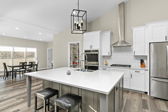 kitchen featuring appliances with stainless steel finishes, white cabinetry, a kitchen island with sink, and wall chimney range hood