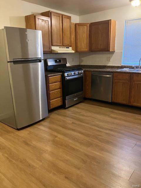 kitchen with light wood-style flooring, dark countertops, under cabinet range hood, appliances with stainless steel finishes, and a sink