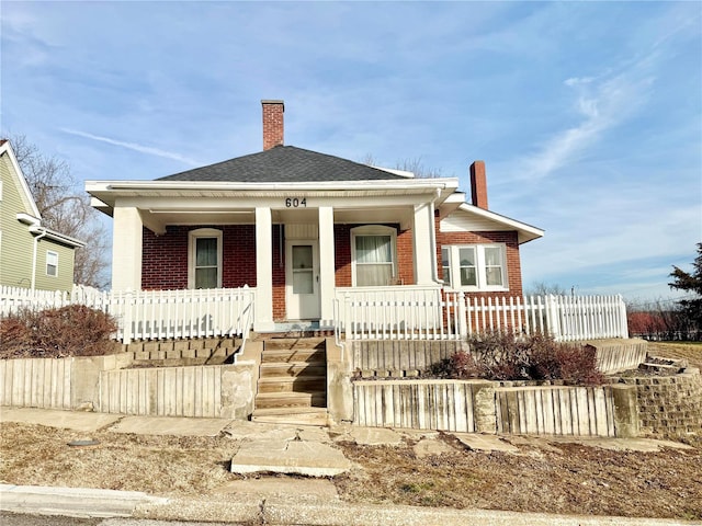 bungalow featuring fence, covered porch, a shingled roof, brick siding, and a chimney