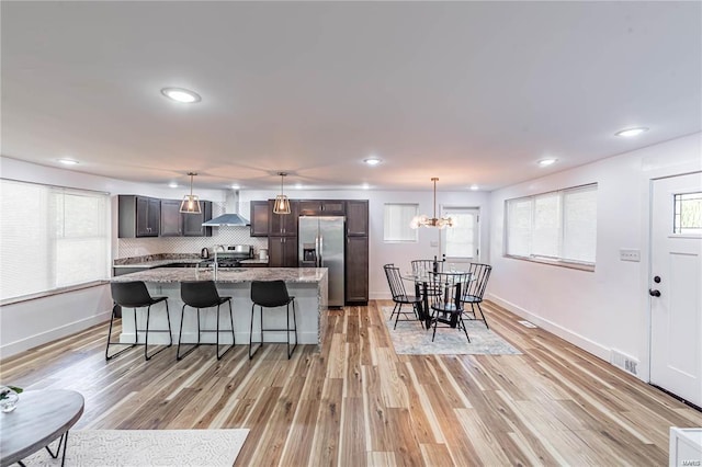 kitchen featuring wall chimney exhaust hood, stainless steel appliances, light hardwood / wood-style flooring, an island with sink, and a chandelier