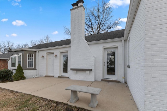 entrance to property featuring a patio, brick siding, roof with shingles, and a chimney