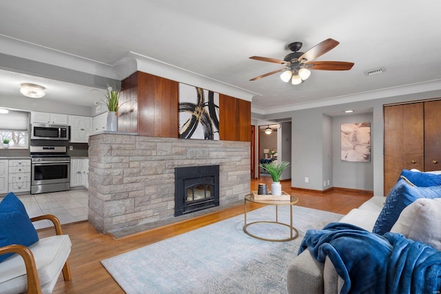 living room featuring crown molding, a stone fireplace, light hardwood / wood-style floors, and ceiling fan