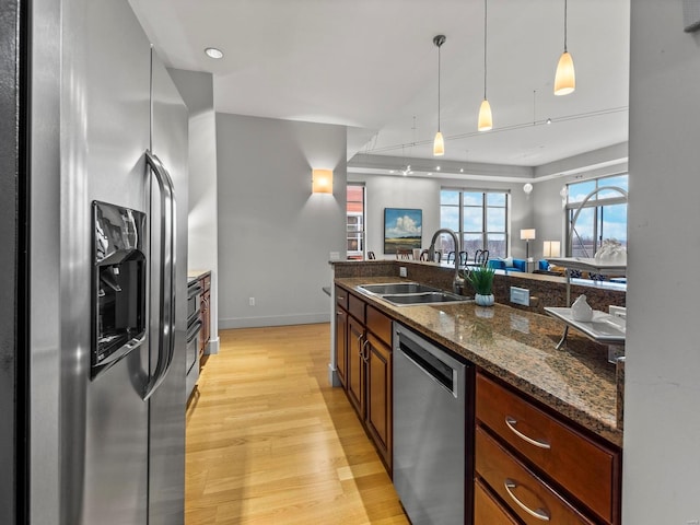 kitchen featuring appliances with stainless steel finishes, light wood-type flooring, decorative light fixtures, sink, and dark stone countertops