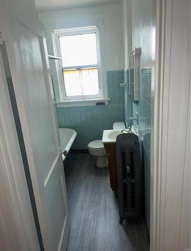 bathroom with a tub to relax in, wood-type flooring, toilet, vanity, and tile walls