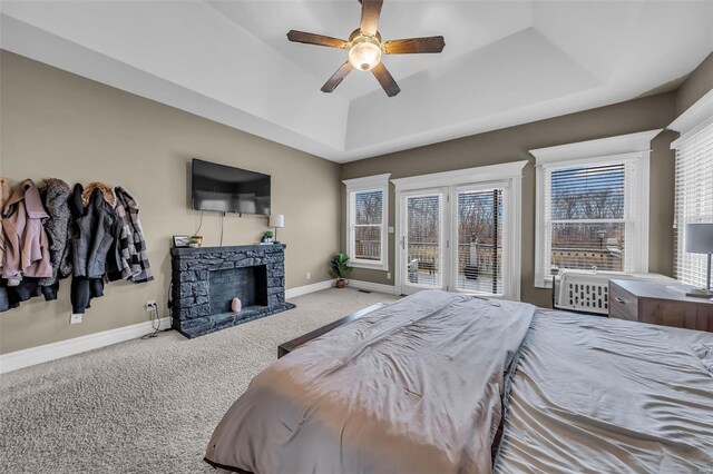 carpeted bedroom featuring a tray ceiling, a ceiling fan, a stone fireplace, access to outside, and baseboards