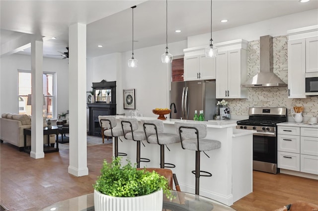 kitchen with wall chimney range hood, backsplash, stainless steel appliances, a center island with sink, and decorative light fixtures