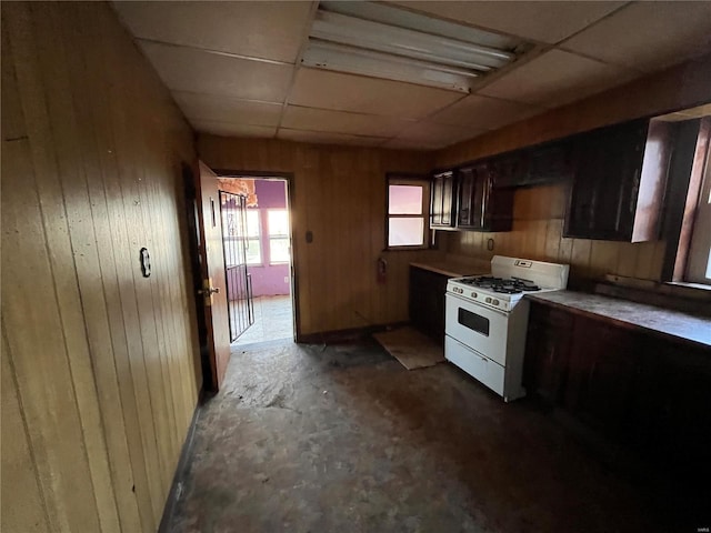 kitchen with a paneled ceiling, white range with gas stovetop, wooden walls, concrete floors, and dark brown cabinetry