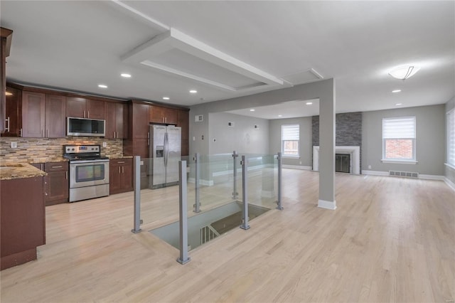 kitchen with appliances with stainless steel finishes, backsplash, light stone counters, light hardwood / wood-style flooring, and a stone fireplace