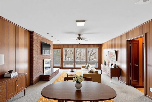 dining area with ceiling fan, wooden walls, a brick fireplace, and light colored carpet