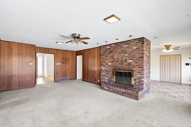 unfurnished living room with visible vents, light colored carpet, ceiling fan, wood walls, and a brick fireplace