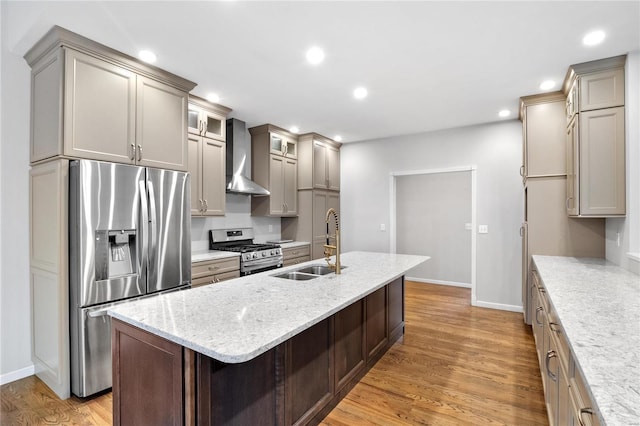 kitchen with sink, wall chimney exhaust hood, light hardwood / wood-style floors, a kitchen island with sink, and appliances with stainless steel finishes