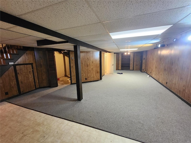 finished basement featuring a drop ceiling, wooden walls, light floors, and stairs