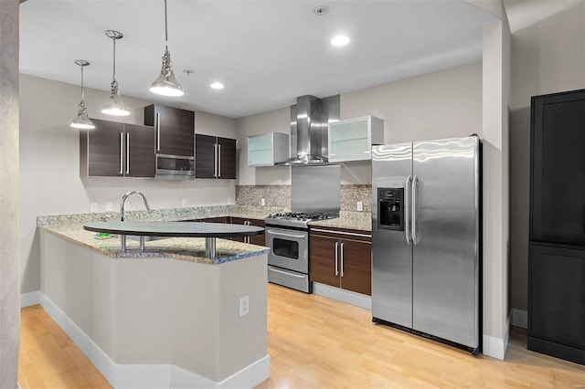 kitchen featuring a peninsula, light wood-style floors, appliances with stainless steel finishes, wall chimney range hood, and light stone countertops