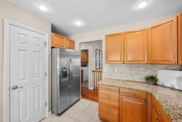 kitchen featuring backsplash, washer / clothes dryer, stainless steel refrigerator with ice dispenser, light tile patterned floors, and light stone counters