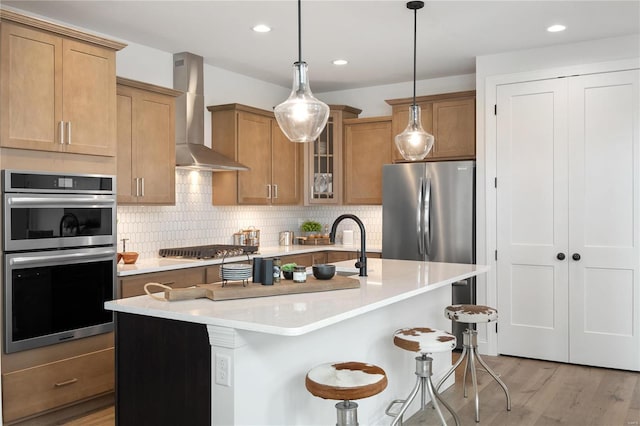 kitchen with a kitchen island with sink, wall chimney exhaust hood, decorative backsplash, light wood-type flooring, and stainless steel appliances