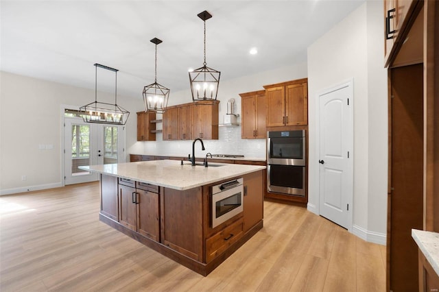 kitchen with light stone counters, multiple ovens, sink, a center island with sink, and hanging light fixtures