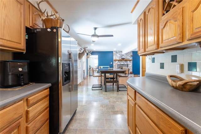 kitchen with decorative backsplash, stainless steel refrigerator with ice dispenser, a brick fireplace, and ceiling fan