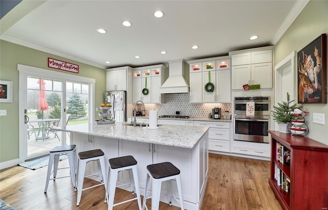 kitchen featuring premium range hood, sink, an island with sink, stainless steel appliances, and light stone counters