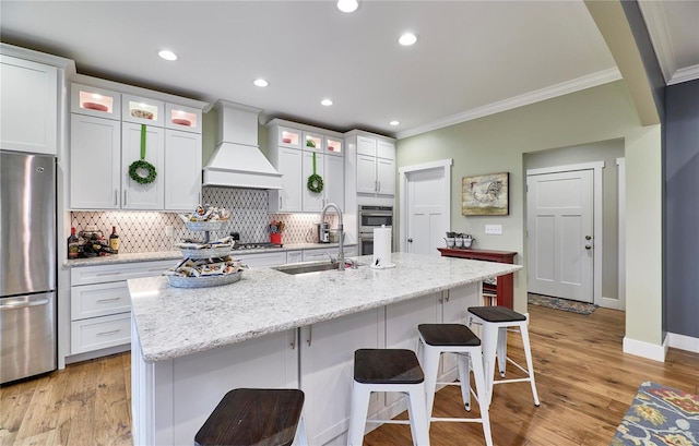 kitchen with sink, custom exhaust hood, white cabinetry, appliances with stainless steel finishes, and a kitchen island with sink