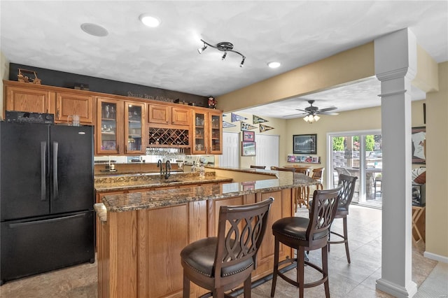 kitchen with ornate columns, ceiling fan, a kitchen breakfast bar, dark stone counters, and black refrigerator