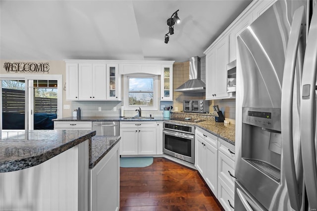 kitchen with a sink, appliances with stainless steel finishes, wall chimney exhaust hood, dark wood finished floors, and glass insert cabinets