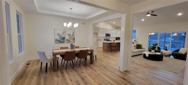 dining room with ceiling fan with notable chandelier, light hardwood / wood-style flooring, and a tray ceiling