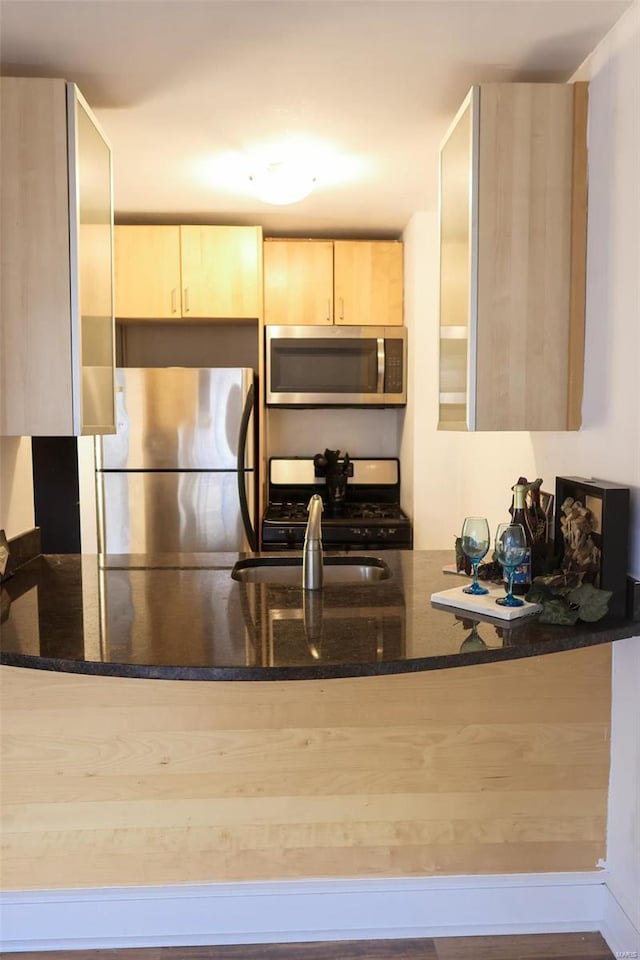 kitchen with kitchen peninsula, sink, stainless steel appliances, light brown cabinetry, and dark stone counters