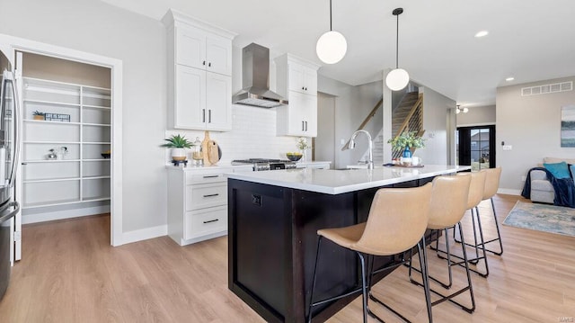 kitchen featuring wall chimney exhaust hood, light hardwood / wood-style flooring, an island with sink, decorative light fixtures, and white cabinets