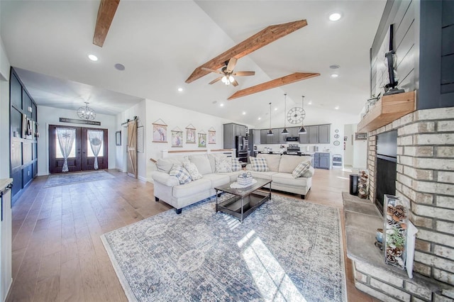 living room featuring ceiling fan with notable chandelier, lofted ceiling with beams, a brick fireplace, light wood-type flooring, and french doors