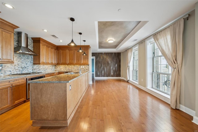 kitchen with dark stone counters, hanging light fixtures, wall chimney exhaust hood, a tray ceiling, and wall oven