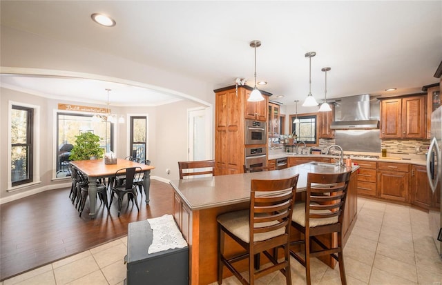 kitchen featuring a kitchen breakfast bar, light tile patterned floors, decorative backsplash, wall chimney range hood, and a kitchen island with sink