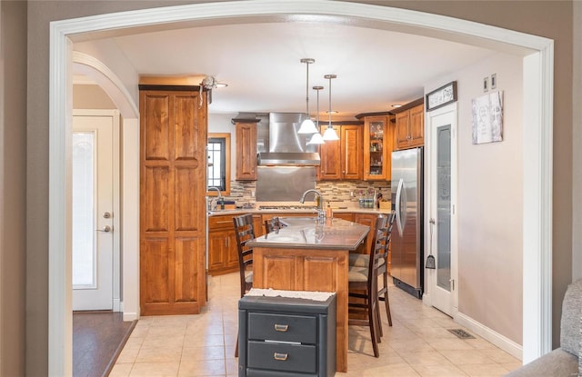 kitchen with an island with sink, exhaust hood, light tile patterned floors, backsplash, and stainless steel refrigerator