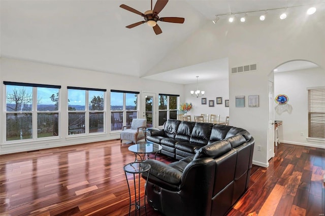 living room featuring rail lighting, a wealth of natural light, ceiling fan with notable chandelier, dark wood-type flooring, and high vaulted ceiling