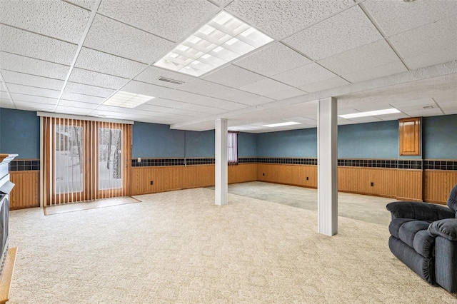 basement featuring a drop ceiling, light colored carpet, and wood walls