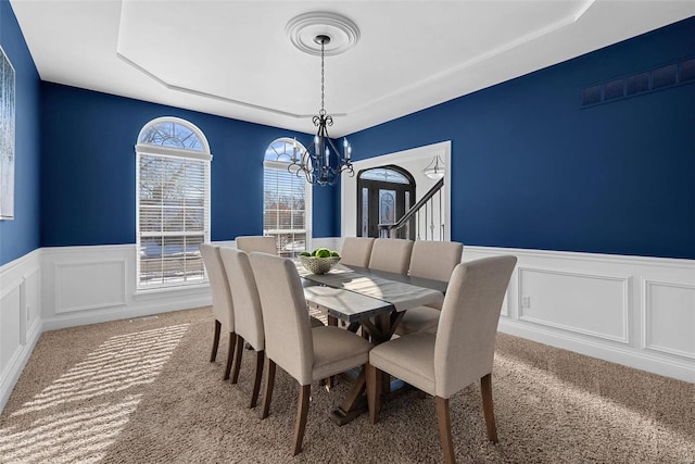 carpeted dining area with visible vents, a notable chandelier, wainscoting, and a decorative wall