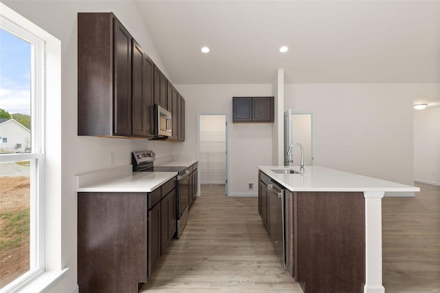 kitchen with appliances with stainless steel finishes, dark brown cabinets, a kitchen island with sink, sink, and light hardwood / wood-style floors