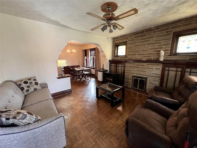 living room with a textured ceiling, ceiling fan with notable chandelier, a stone fireplace, and dark parquet floors